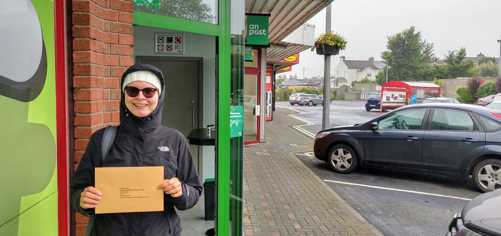 Kobieta w kurtce przeciwdeszczowej i czapce stoi przed budynkiem poczty, trzymając w ręce kopertę z kartą do głosowania.
